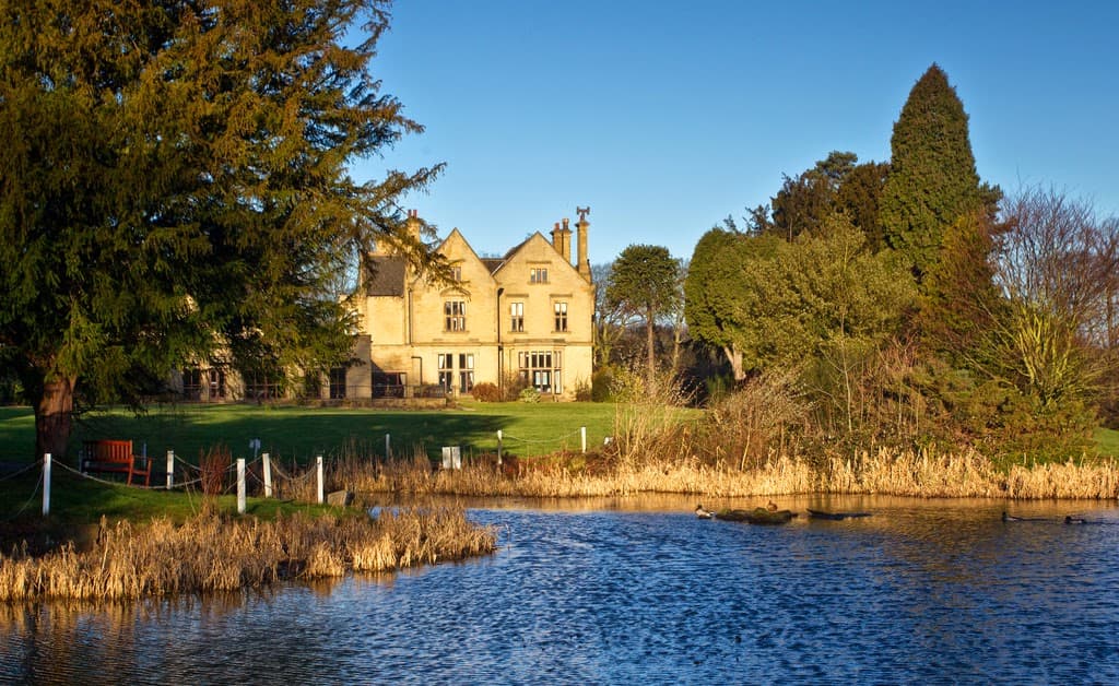 Historic Bagden Hall Hotel beside a tranquil lake, surrounded by greenery and trees under a clear blue sky.