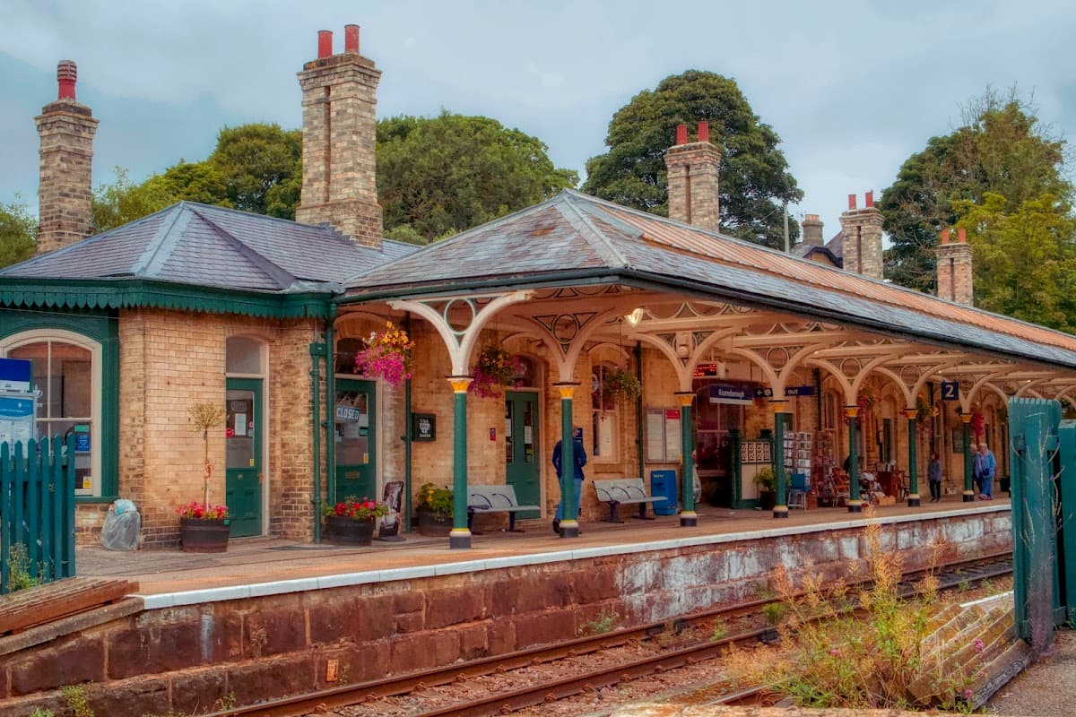 Victorian-style railway station with brick buildings, covered platforms, and potted flowers, surrounded by trees.