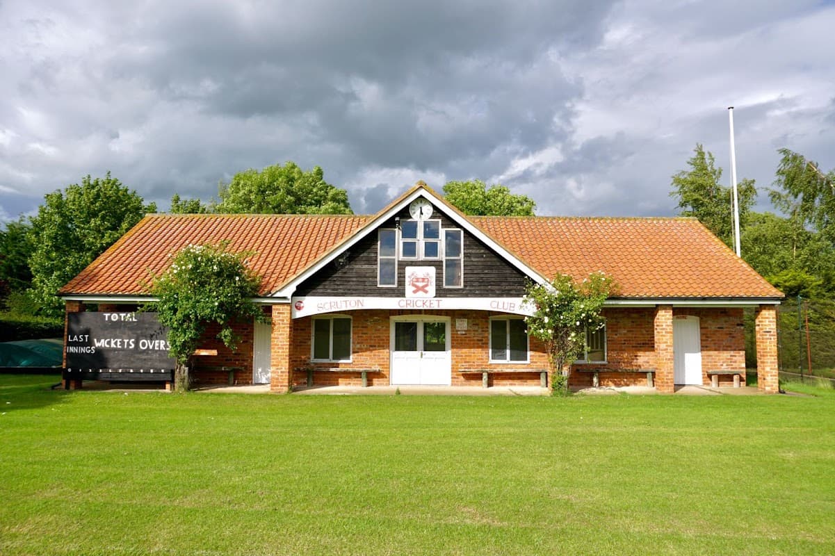Scruton Cricket Club building with a red-tiled roof, green lawn, and cloudy sky, featuring a sign and scoreboard.