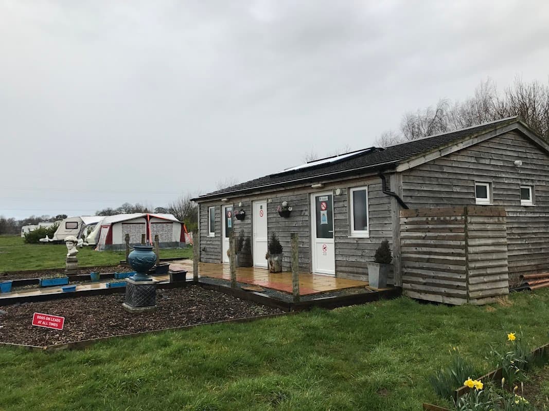 Wooden lodge with two doors, garden area, and a fountain, surrounded by grassy fields and a tent in the background.