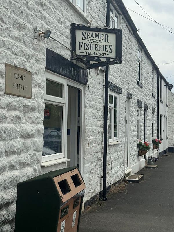 White stone building with a sign reading "Seamer Fisheries" and flower boxes under the windows. Litter bins nearby.