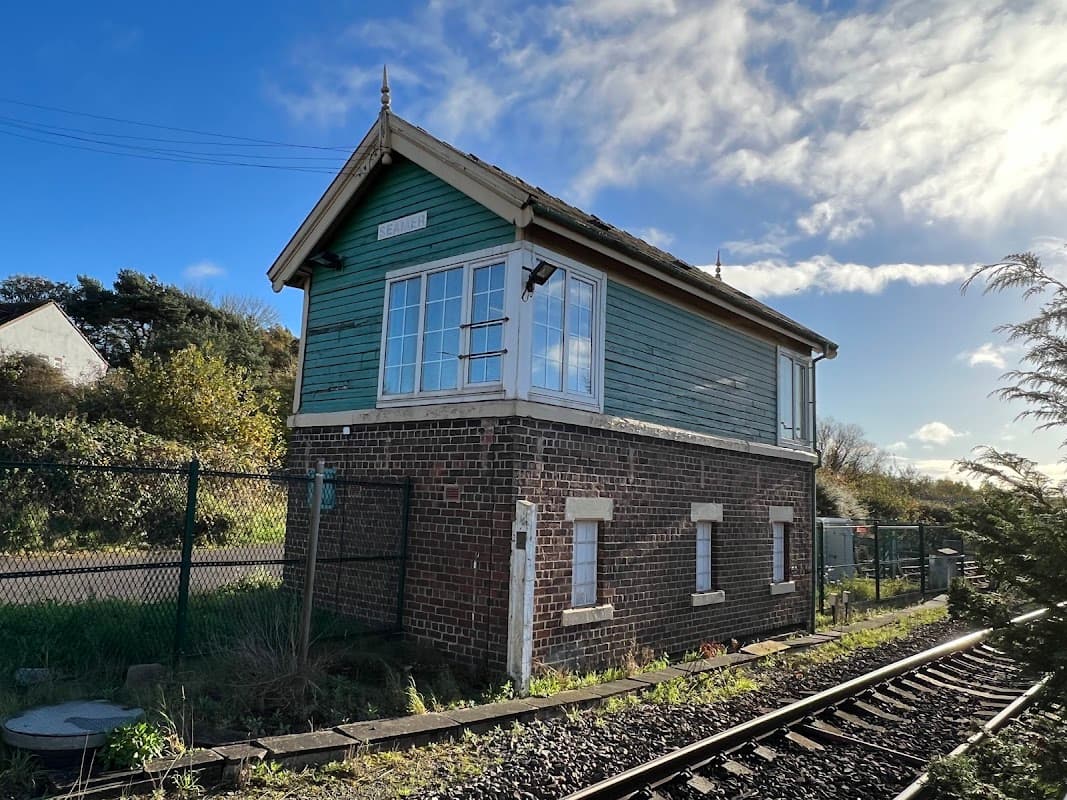 Historic railway signal box with green wooden exterior, surrounded by greenery and tracks under a blue sky.