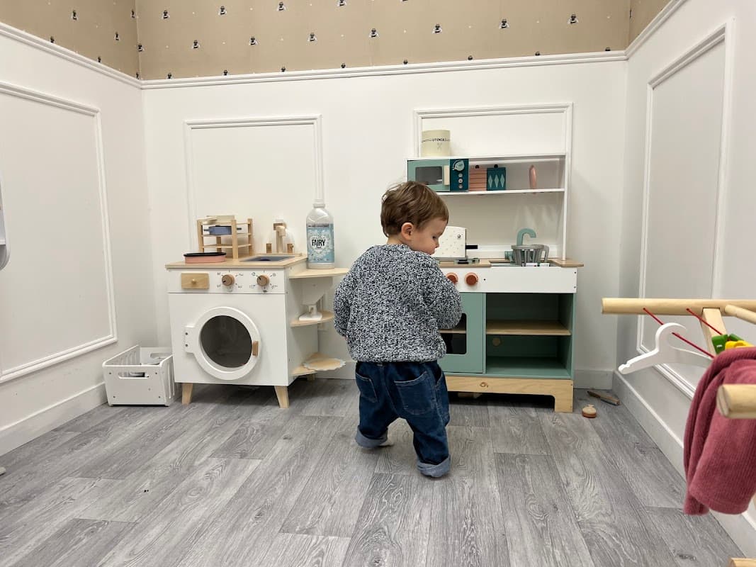 A toddler playing in a miniature kitchen with a toy stove, sink, and washing machine in a bright, playful room.