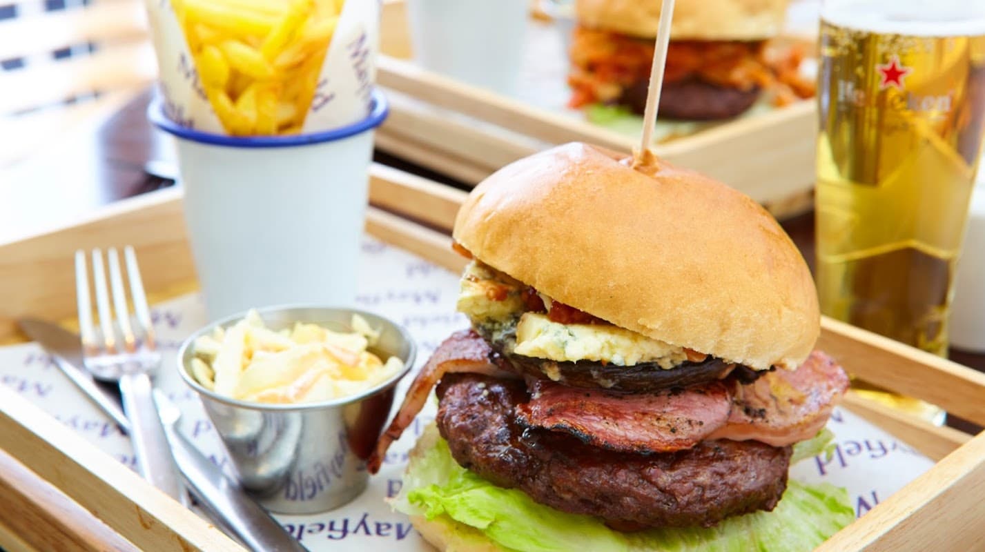 Juicy burger with bacon, cheese, and lettuce, served with fries and coleslaw on a wooden tray. Beer in the background.