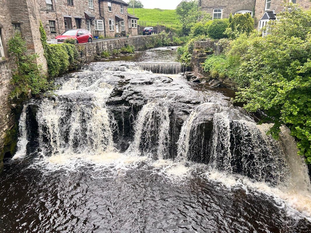 A picturesque cottage by a flowing stream with small waterfalls, surrounded by lush greenery in Sedbusk, Yorkshire.