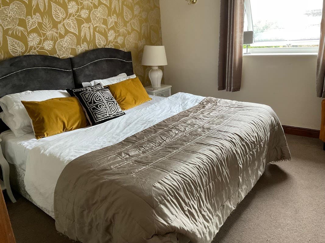 Cozy bedroom with a large bed, decorative pillows, bedside lamp, and patterned wallpaper in a cottage setting.