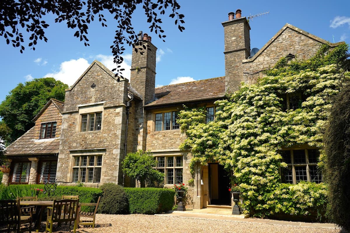 Historic stone building with ivy-covered walls, surrounded by greenery and outdoor seating in a tranquil setting.
