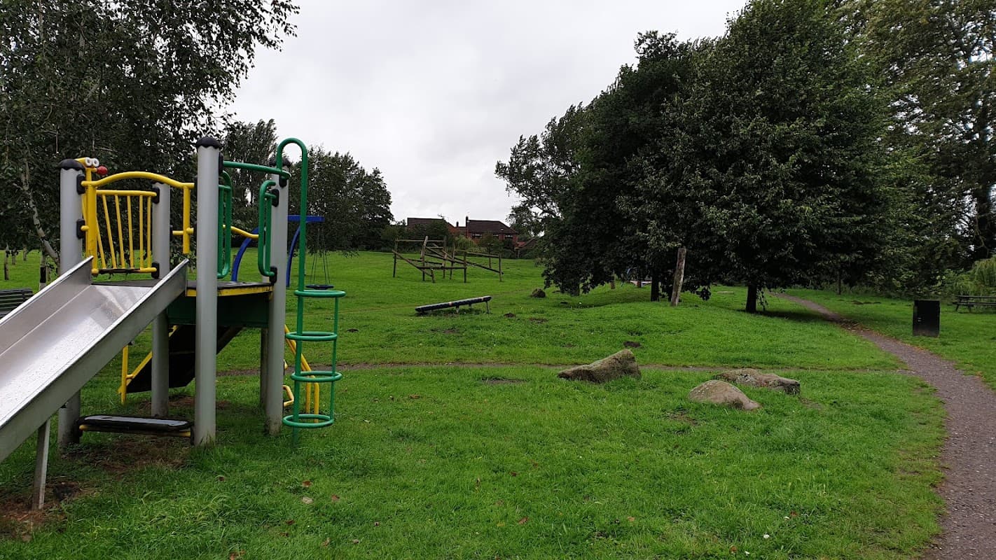 Playground slide and climbing structure in a grassy park with trees and a path leading through the area.