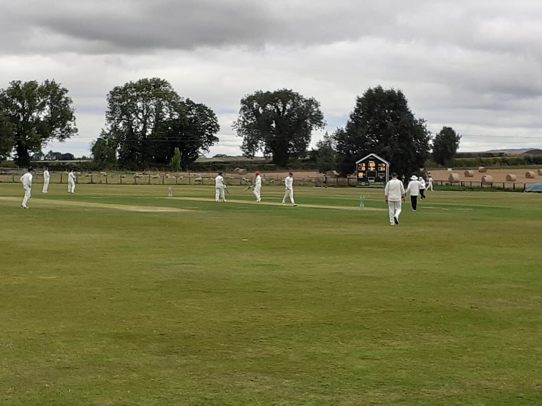 Cricket match in progress at Sessay Cricket Club, with players in white uniforms and a scoreboard in the background.