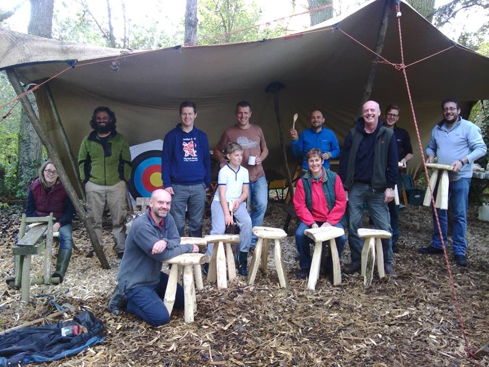 Group of people posing under a large tarp with wooden stools and an archery target in a woodland setting.