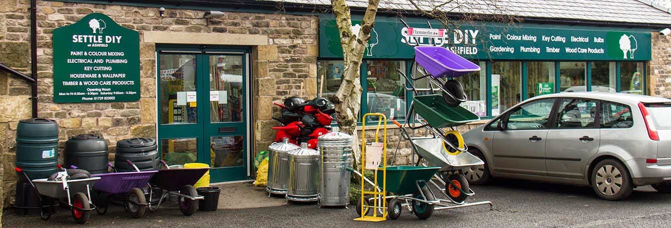 Colorful garden carts and tools are displayed outside Settle DIY shop, with a parked car nearby.