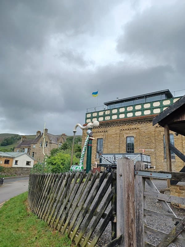 Car park with stone buildings, a flag, and a wooden fence under a cloudy sky in Settle, Yorkshire.