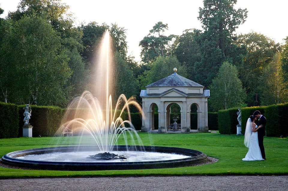 A couple embraces near a fountain in a landscaped garden, with a classical pavilion and trees in the background.