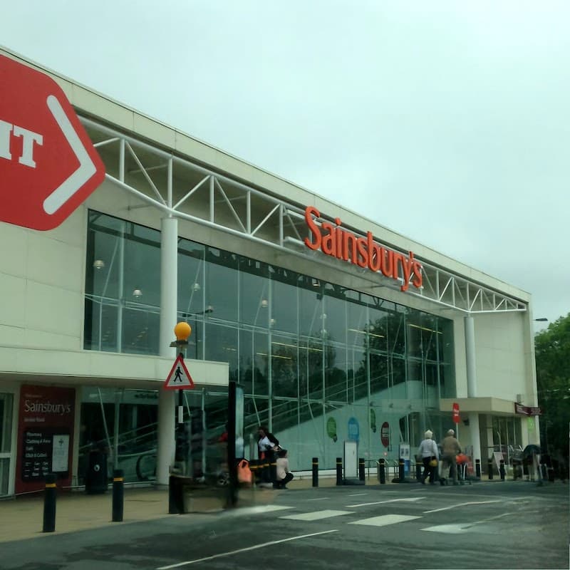 Sainsbury's storefront with large glass windows, red signage, and a few people outside under a cloudy sky.