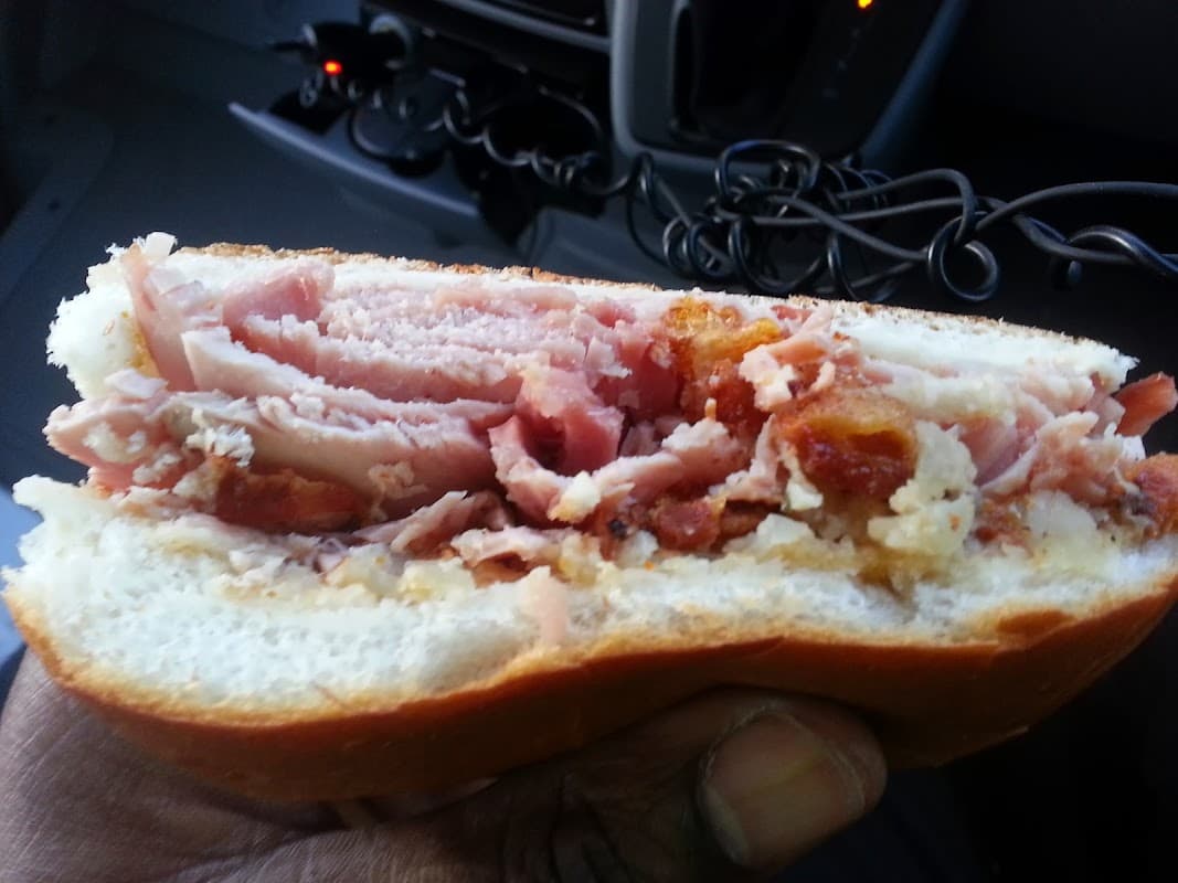 Close-up of a sandwich filled with layers of ham and crispy bacon, held in a hand, with a vehicle interior in the background.