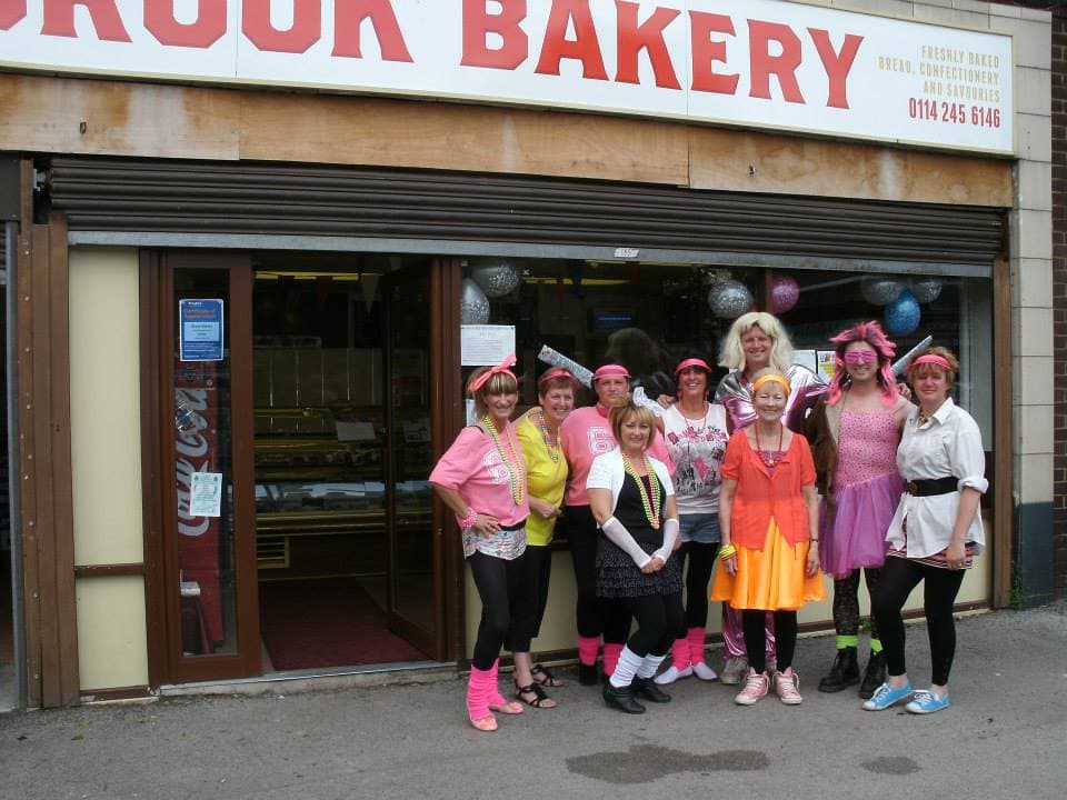 A group of cheerful bakers in colorful outfits stands outside Brook Bakery Ltd, with balloons and a welcoming storefront.