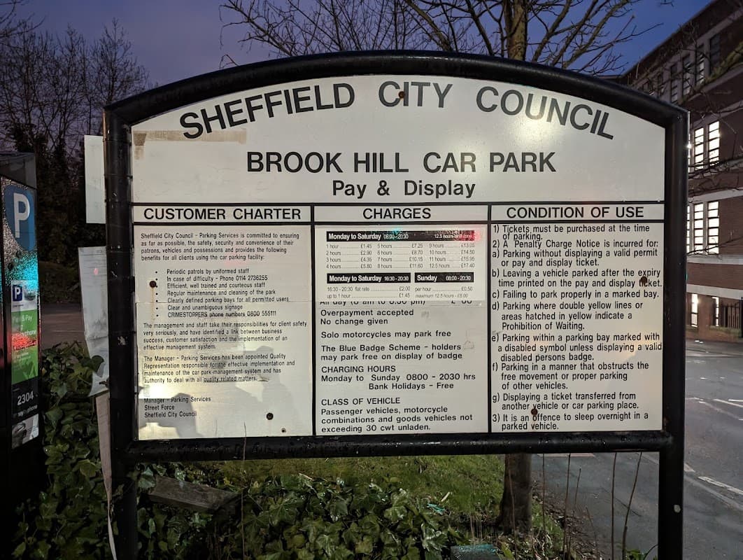 Sign for Brook Hill Car Park detailing customer charges, conditions, and operational hours, set against a dimly lit background.
