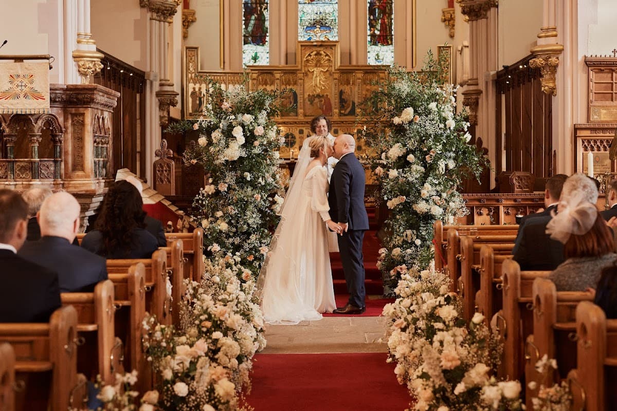 A couple exchanges vows in a decorated church, surrounded by floral arrangements and seated guests.
