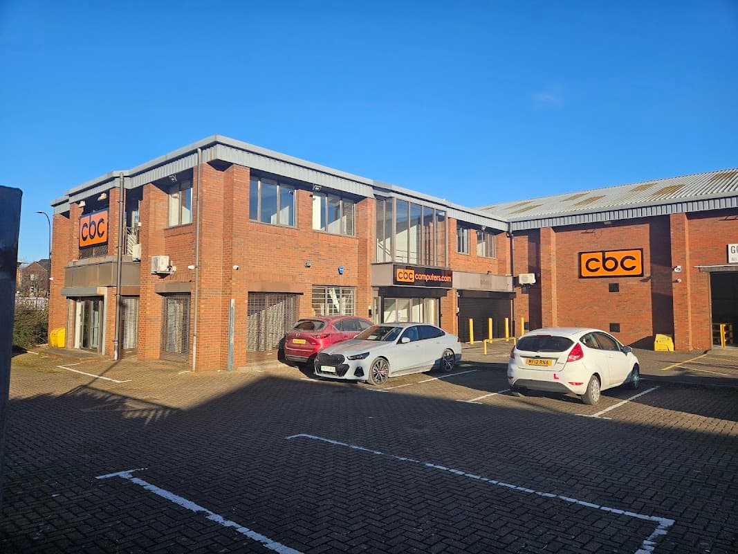 Brick building with large windows and "cbc" signage, surrounded by parked cars in a paved lot under a clear blue sky.