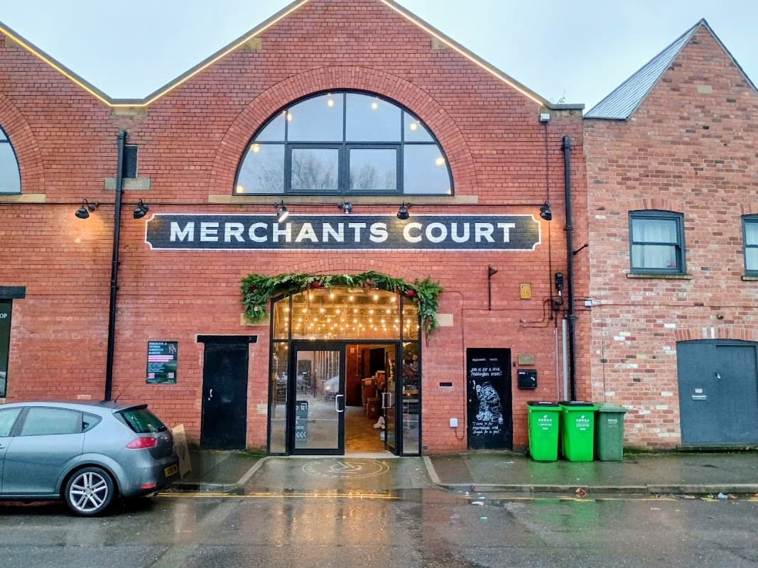 Brick building entrance with "MERCHANTS COURT" sign, festive lights, and green bins nearby. Wet pavement reflects light.