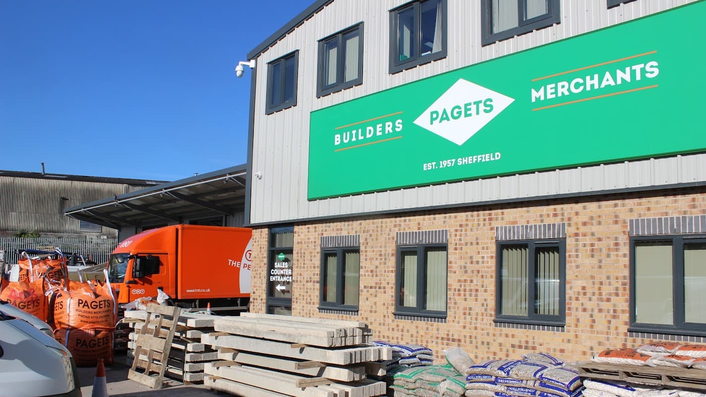 Bright green sign reading "Pagets Builders Merchants" on a brick building, with orange delivery truck and stacked materials outside.