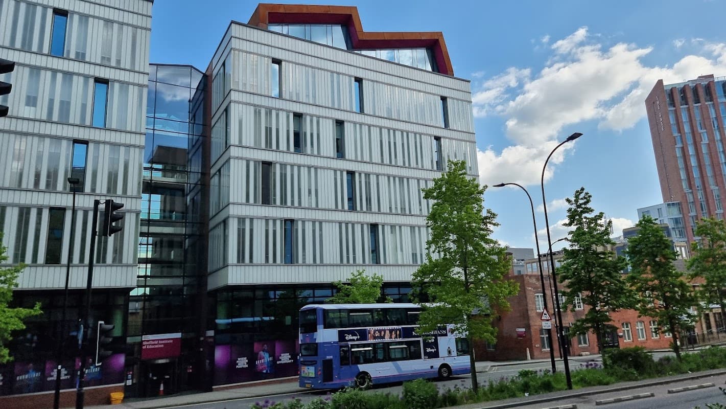 Modern building with glass facade, trees lining the street, and a bus stopped in front on a sunny day.