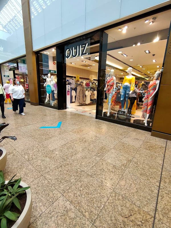 Storefront of Quiz Clothing featuring mannequins in colorful outfits, shoppers, and a modern interior in a mall setting.