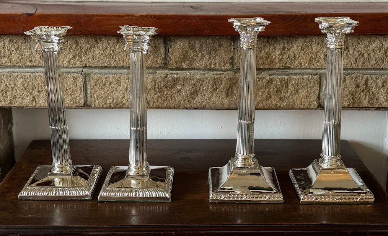 Four ornate silver candlesticks with fluted columns and square bases, displayed on a wooden surface against a stone wall.