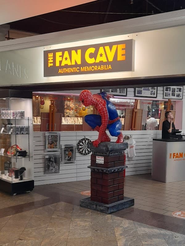 A colorful Spider-Man statue crouches on a brick pedestal in front of "The Fan Cave" shop, featuring memorabilia displays.