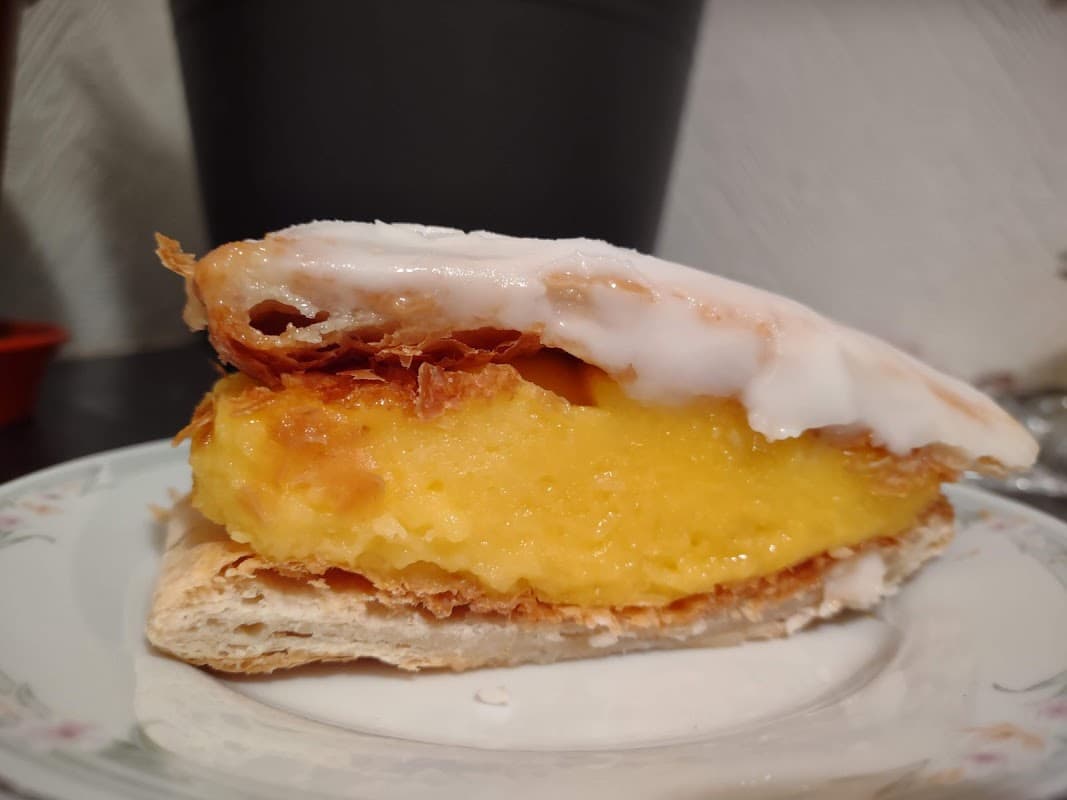A slice of pastry topped with icing, revealing a creamy yellow filling, served on a decorative plate.