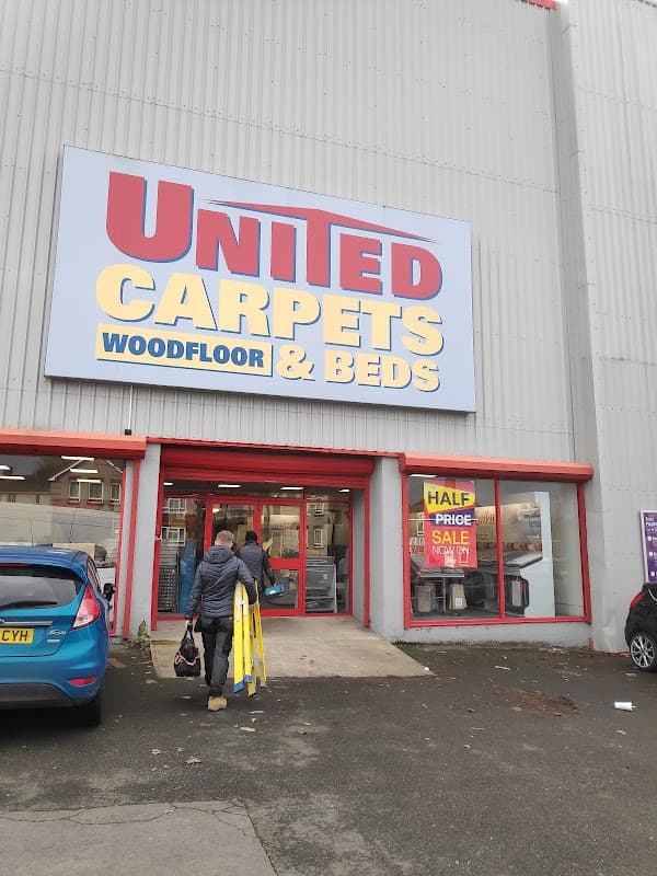 Storefront of United Carpets and Beds with a large sign, a person carrying a ladder, and a parked car.