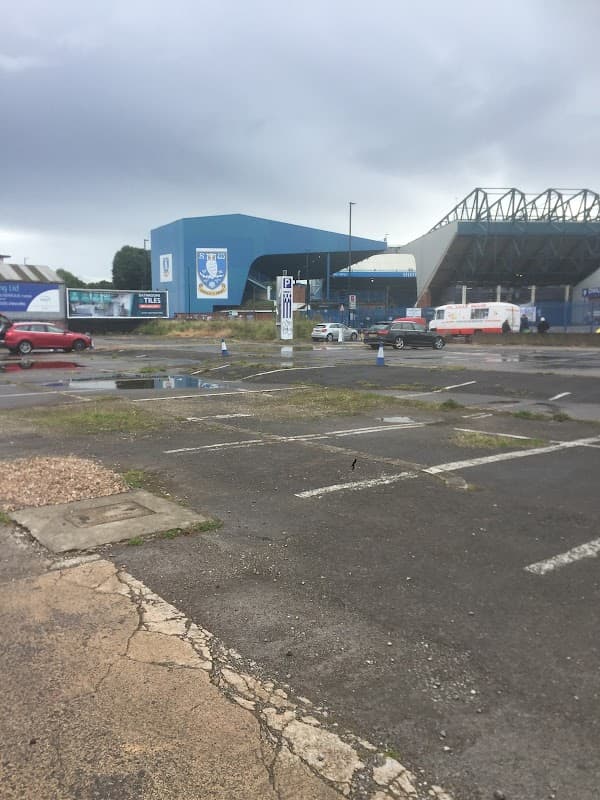 Empty car park with puddles, featuring a blue building and Sheffield Wednesday signage in the background.