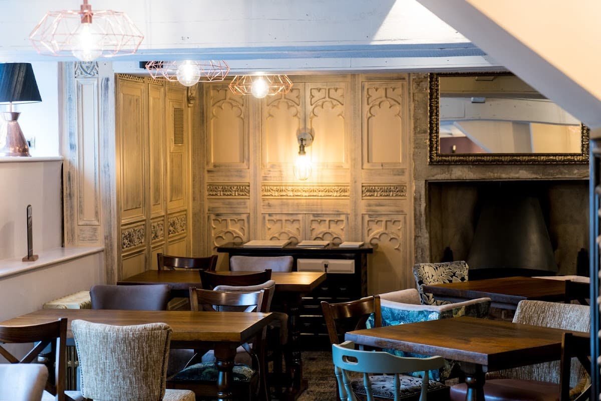 Cozy bar interior with wooden tables, upholstered chairs, ornate wall paneling, and warm lighting fixtures.