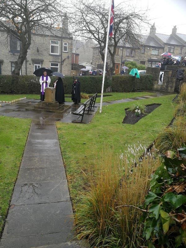 Memorial Gardens in Shelf, Yorkshire, featuring a stone monument, benches, and people under umbrellas in rainy weather.