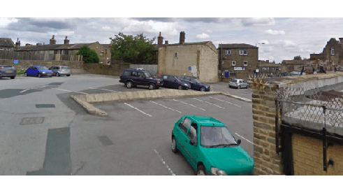 Car park with multiple vehicles, including a green car, surrounded by buildings in Shelf, Yorkshire.