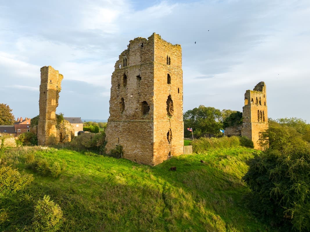 Ruins of Sheriff Hutton Castle with grassy mounds and trees, under a blue sky with scattered clouds.