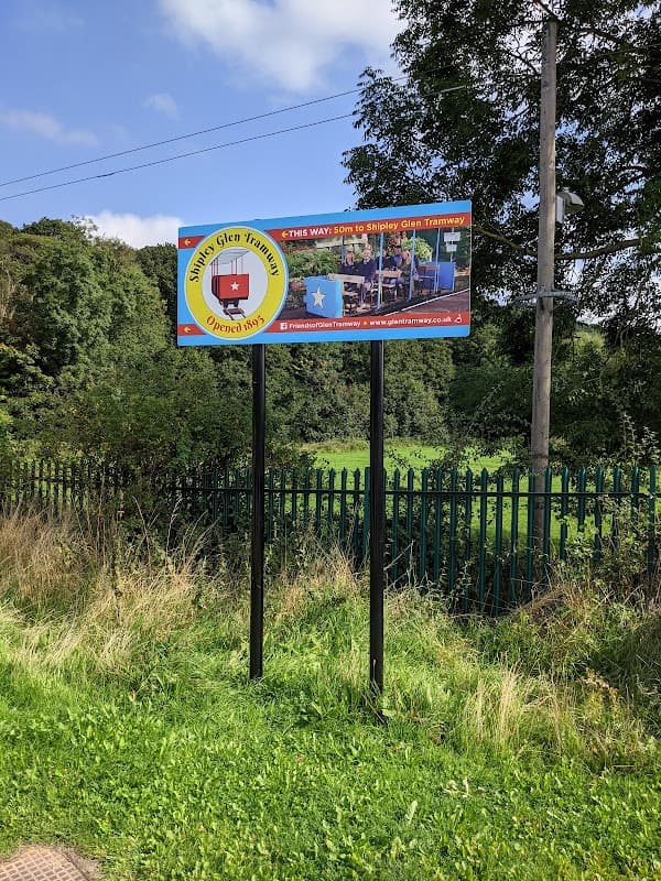 Sign for Shipley Glen Tramway with images and directions, surrounded by greenery and a fence.