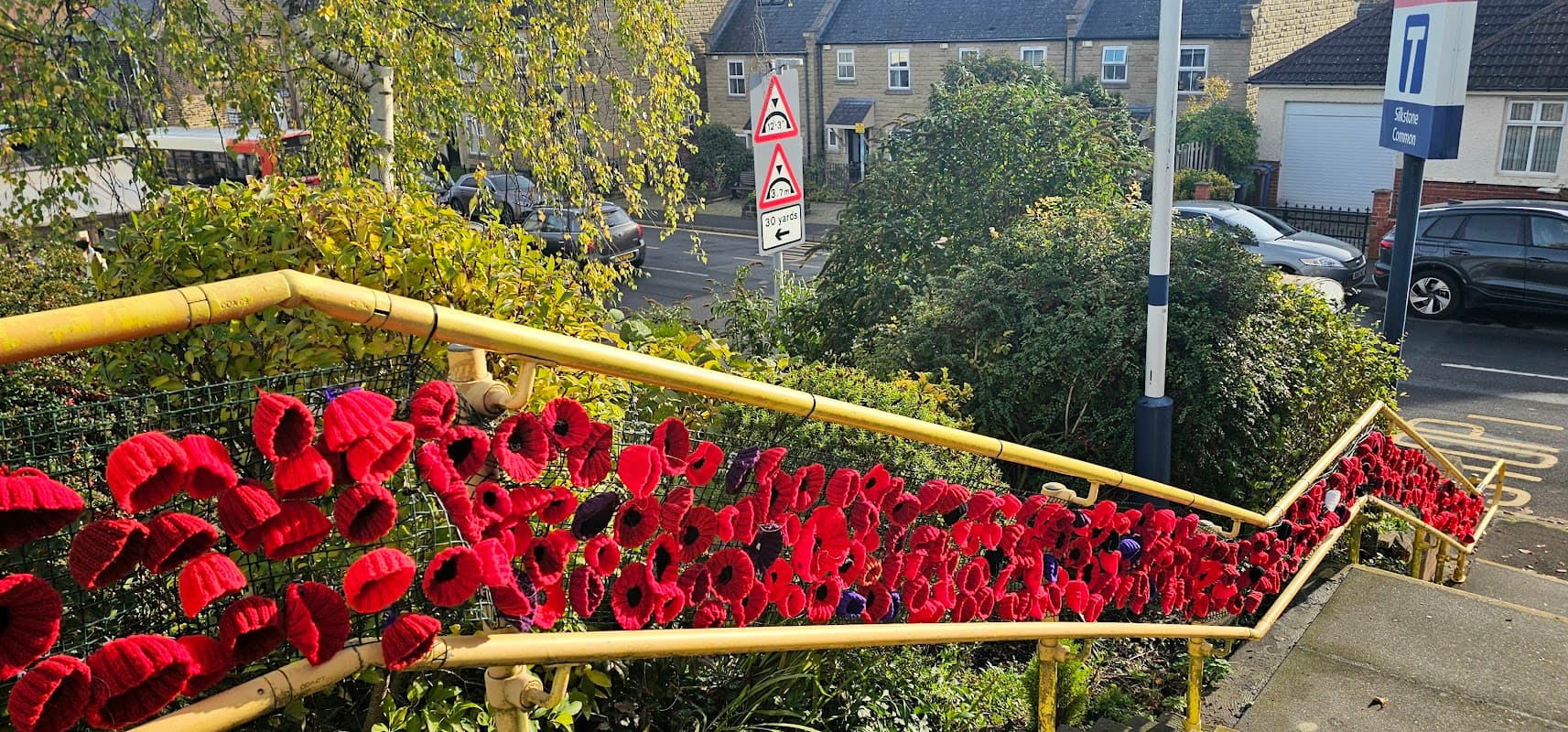 Colorful knitted poppies adorn a yellow railing along a staircase, with cars and houses in the background.