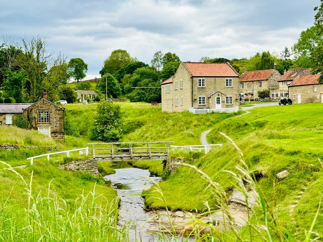 Quaint stone buildings with red roofs by a stream, surrounded by lush green fields and trees in Sinnington, Yorkshire.
