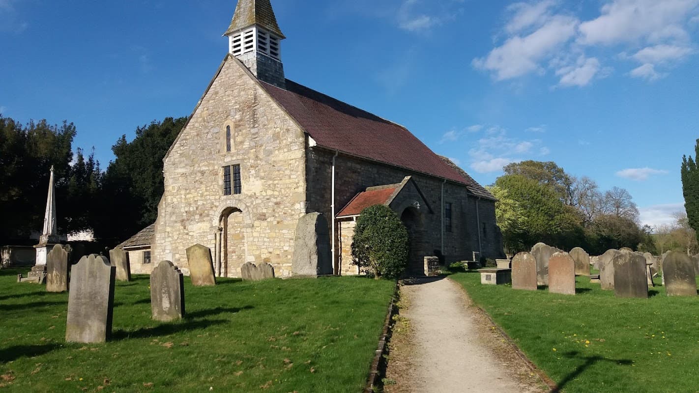 Stone church with a bell tower surrounded by gravestones and green grass under a blue sky with scattered clouds.