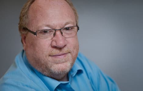 A middle-aged man with glasses and a beard, wearing a light blue shirt, smiles confidently at the camera.