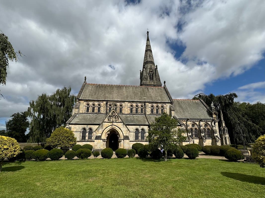 Victorian church with a tall spire, surrounded by manicured lawns and trees under a partly cloudy sky.