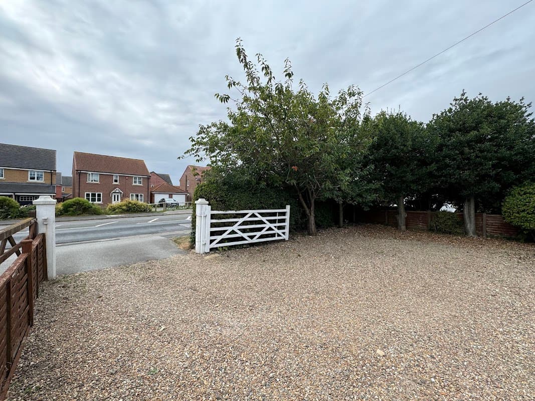 Gravel parking area with a white gate, surrounded by trees and houses under a cloudy sky in Skidby, Yorkshire.
