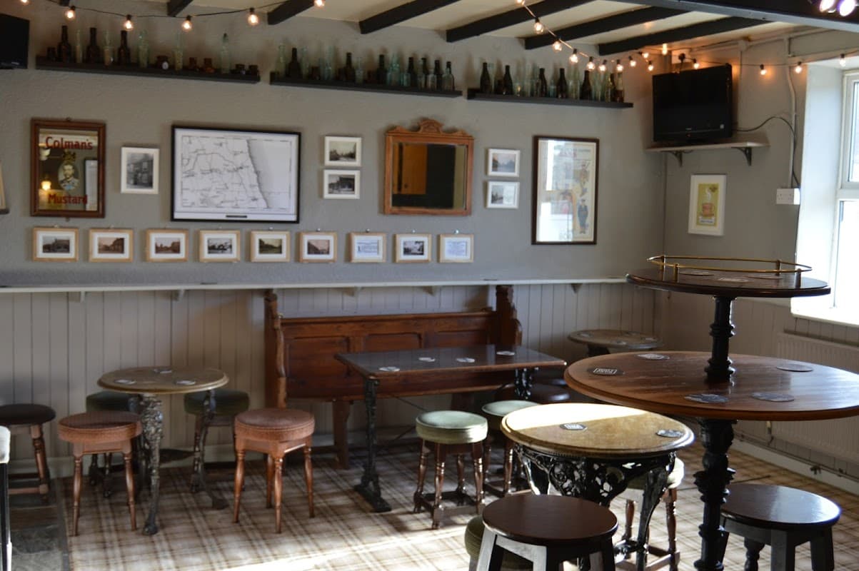 Cozy bar interior with wooden tables, stools, framed photos, and vintage bottles along the walls, illuminated by string lights.