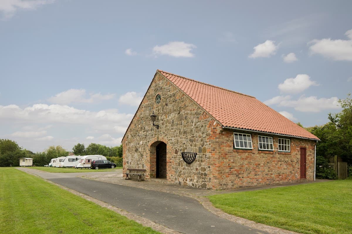 Stone building with a red-tiled roof, surrounded by green grass and parked caravans under a cloudy sky.