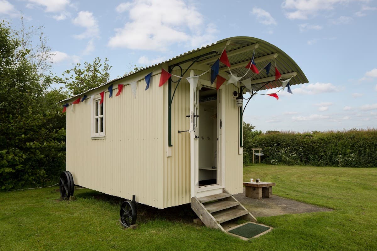 A charming shepherd's hut with a curved roof, bunting, and steps, set in a grassy area with trees in the background.