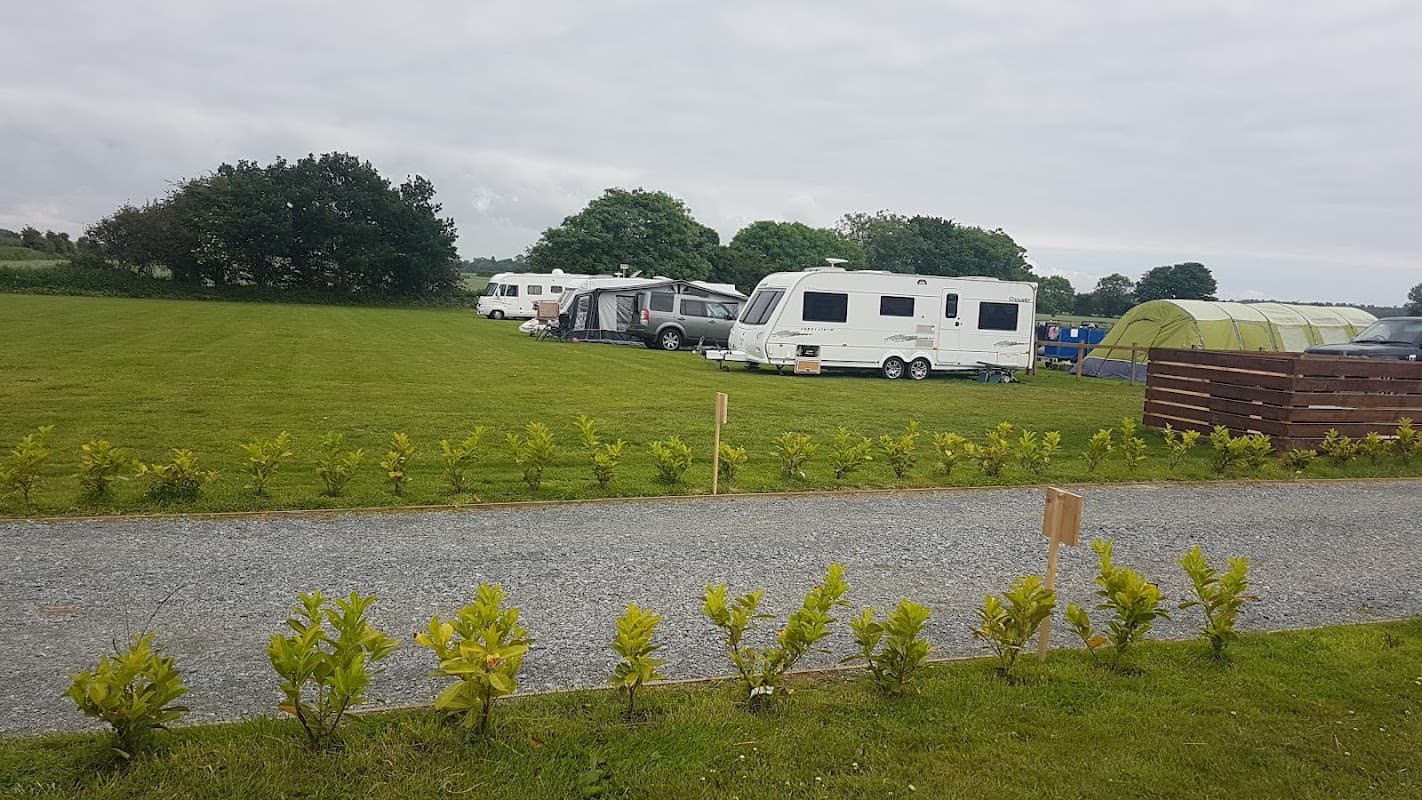 Caravans parked on a grassy field, bordered by a neat hedge, with trees and camping tents in the background.