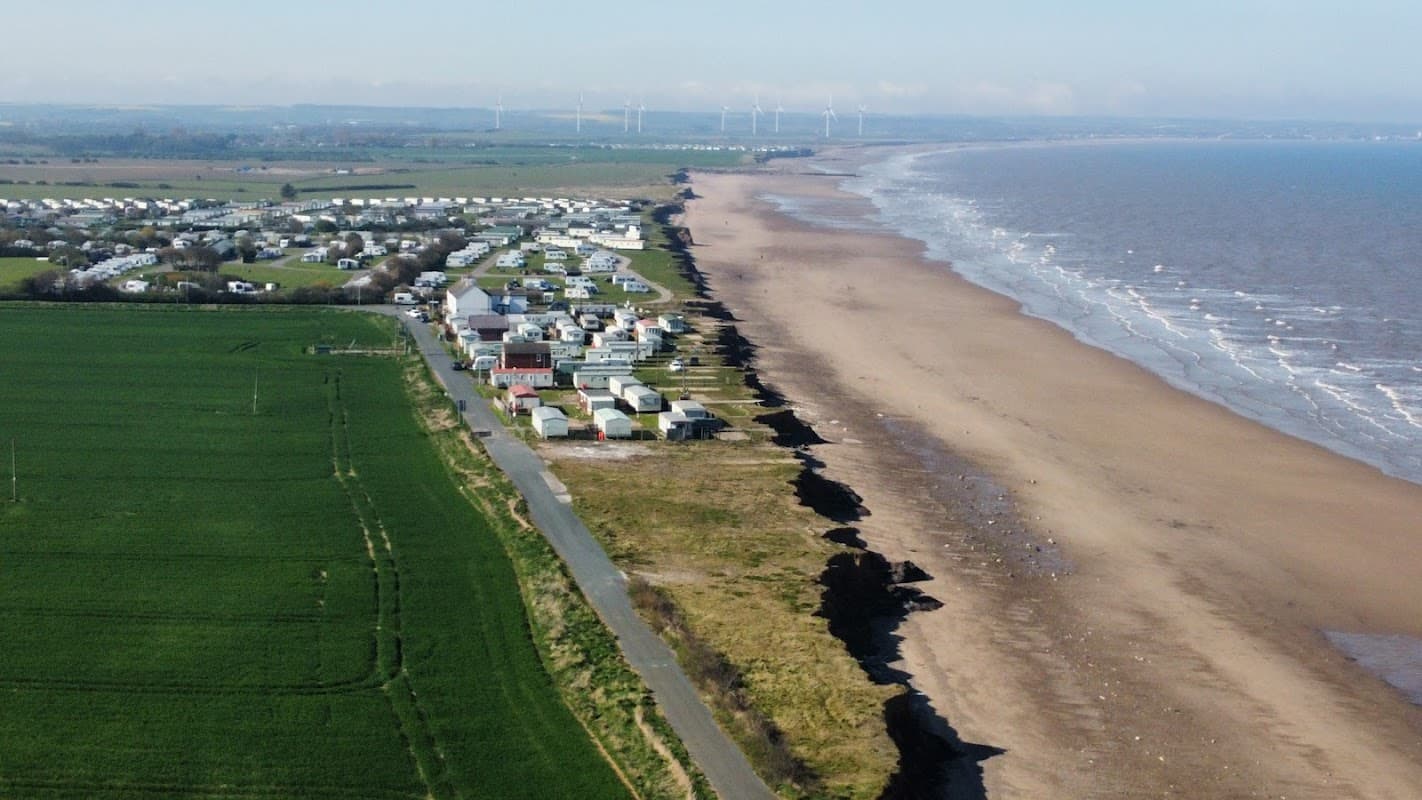 Coastal view of Skipsea Sands with caravan park, green fields, and sandy beach alongside the sea. Wind turbines in the distance.