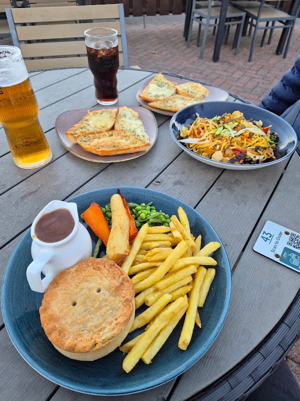 A table set with a pie, fries, peas, carrots, and salad, alongside drinks and garlic bread in a casual cafe setting.