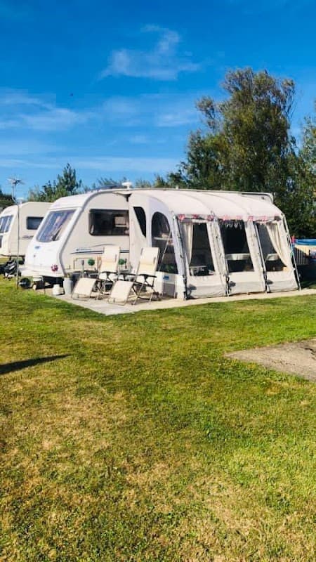 Caravan with an awning and outdoor chairs on a grassy area, surrounded by trees under a clear blue sky.